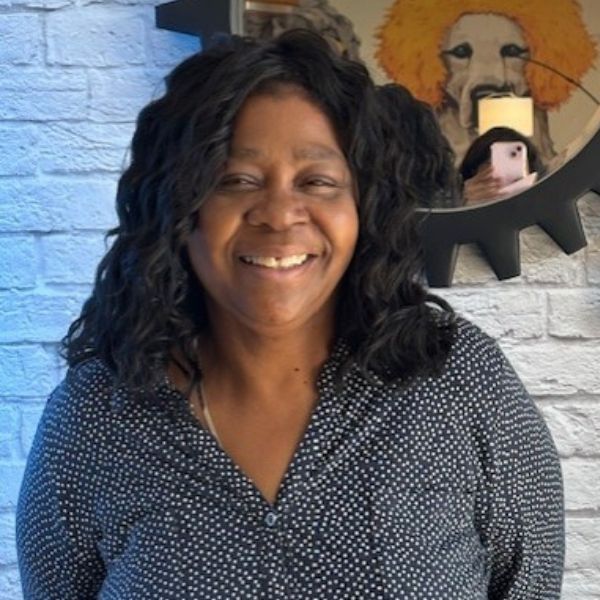 AR Kids Read (1) A woman with wavy black hair wearing a dark blue polka-dotted shirt smiles in front of a white brick wall. A round mirror behind her reflects a person taking a photo and a painting of a lion.
