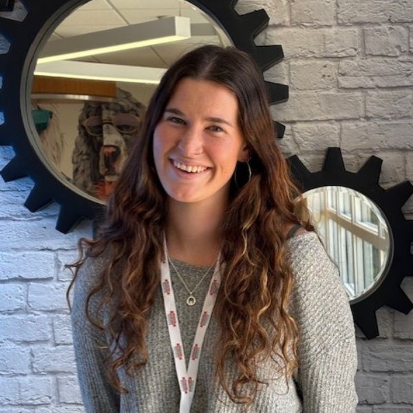 AR Kids Read (10) A young woman with long wavy brown hair, wearing a gray sweater and a lanyard, smiles in front of a gray brick wall decorated with large black gear-shaped wall pieces.