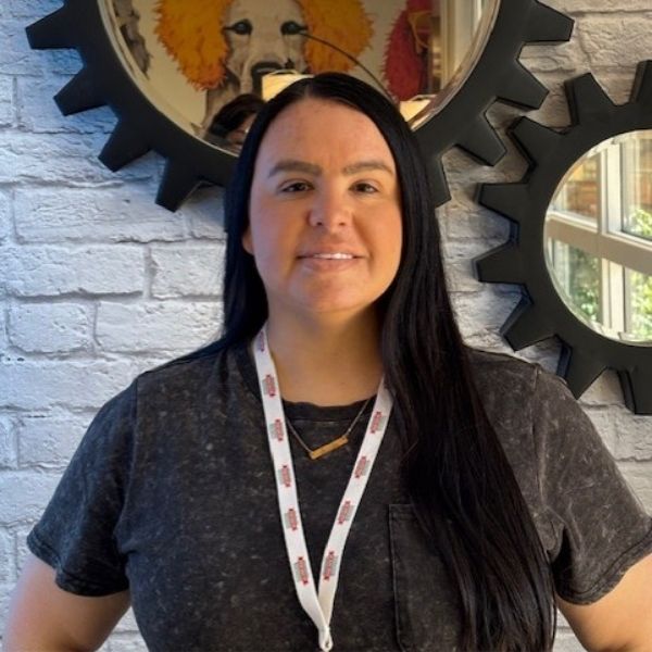AR Kids Read (2) A woman with long black hair, wearing a dark t-shirt and a lanyard, stands in front of a white brick wall with circular mirrors and a colorful artwork reflected in one of the mirrors.