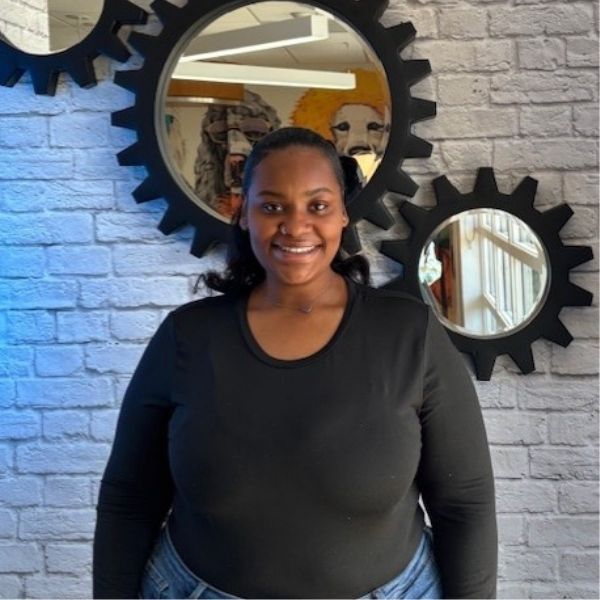 AR Kids Read (3) A woman wearing a black top and blue jeans smiles at the camera, standing in front of a white brick wall decorated with round mirrors framed by black gear shapes.