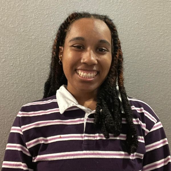 AR Kids Read (4) A person with long twisted hair smiles at the camera. They are wearing a purple and white striped collared shirt and standing in front of a plain gray wall.
