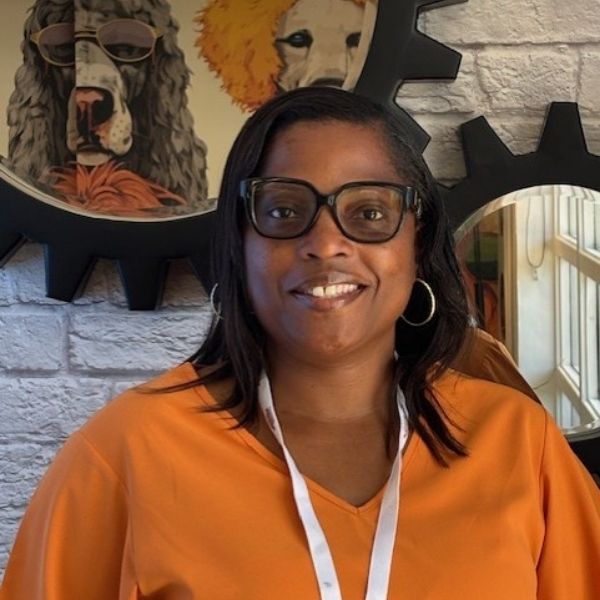 AR Kids Read (6) A woman with glasses and a white lanyard, wearing an orange shirt, smiles in front of a white brick wall decorated with framed animal artwork and gear-shaped mirrors.