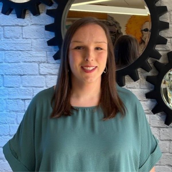 AR Kids Read (7) A woman with straight brown hair, wearing a teal top, stands smiling in front of a white brick wall decorated with black gear-shaped mirrors.