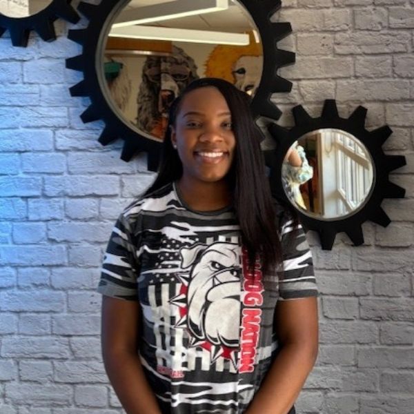 AR Kids Read (8) A smiling woman with straight, long hair stands in front of a white brick wall with gear-shaped mirrors. She wears a black and white Bulldog Nation t-shirt with a large bulldog graphic.