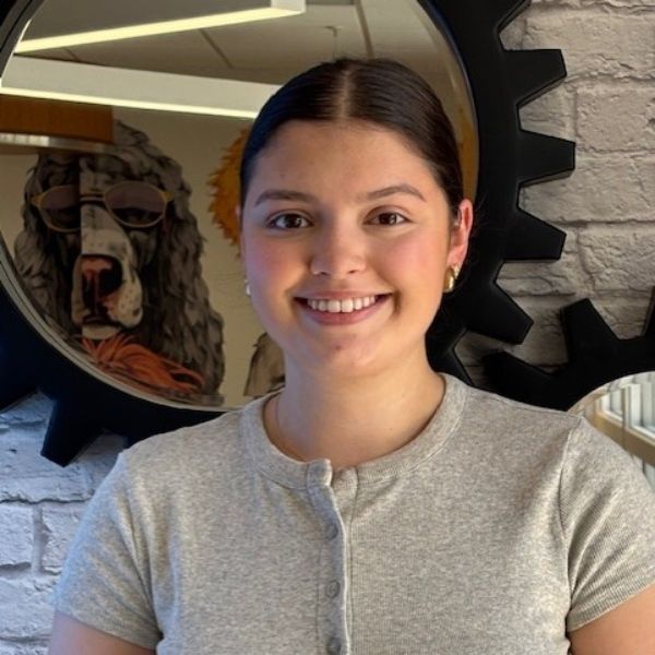 AR Kids Read (9) A smiling young woman with dark hair in a bun, wearing a light gray button-up top, stands indoors in front of a round mirror and brick wall, with a dog statue in sunglasses reflected behind her.