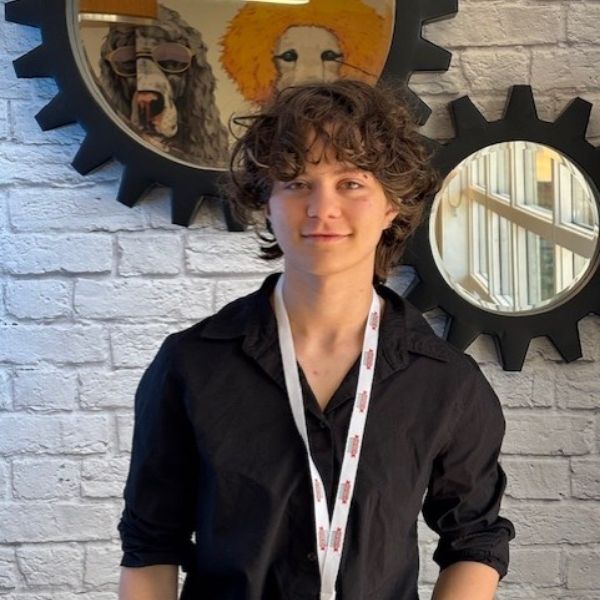 AR Kids Read A young person with curly hair wearing a black shirt and a white lanyard stands in front of a white brick wall decorated with round, gear-shaped frames, one featuring artwork of dogs in wigs and glasses.