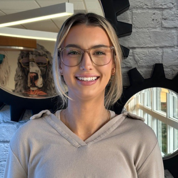 McKenna Knott-Debord A smiling woman with glasses and a beige hoodie stands in front of a white brick wall and a round mirror with a gear-shaped frame. A reflection behind her shows a dog wearing sunglasses.