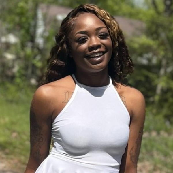 Melissa Stone A woman with long wavy hair wearing a sleeveless white halter-top dress smiles outdoors with greenery and a house blurred in the background.