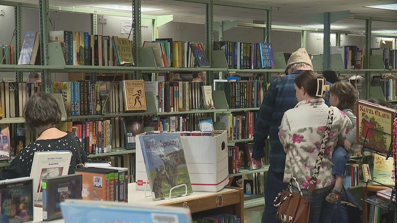 People browse books in a library or bookstore, with shelves filled with colorful book spines. Some are reading, while others are standing and looking at the shelves. The scene is indoors and well-lit.