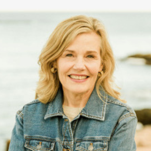 A smiling woman with blonde hair wearing a denim jacket stands outdoors near the coast, with the ocean and rocks visible in the background.