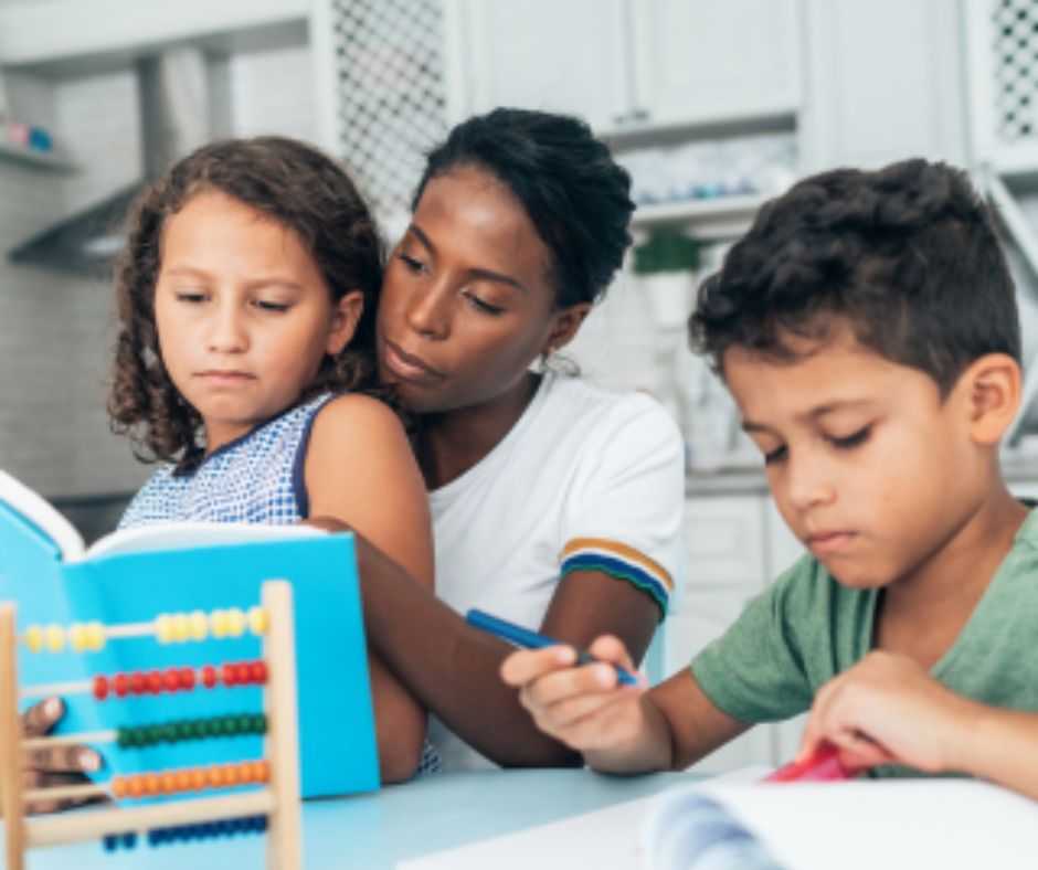 A woman helps a young girl read a book while a boy writes in a notebook beside them. An abacus sits on the table in the foreground. The scene takes place in a bright, modern kitchen.