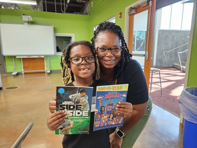 A smiling child and an adult woman stand indoors, holding up two books: one titled Side by Side: Baseball Stars and the other Harlem Globetrotters: Ready-to-Read. They appear happy and proud.