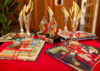 A round red table displays several gold and glass trophies along with colorful childrens books tied with red ribbon, suggesting a prize or award ceremony setting.