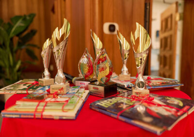 A group of trophies and colorful wrapped books are displayed on a red tablecloth, with a wooden wall and green plant in the background.