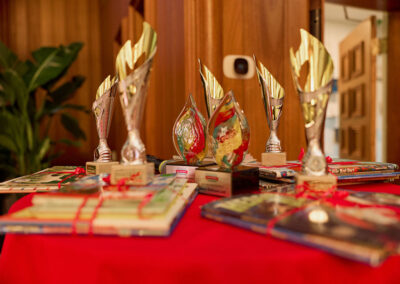 A red tablecloth covers a round table displaying several trophies and plaques, with stacks of books tied with red string, set indoors near wooden decor and green plants.