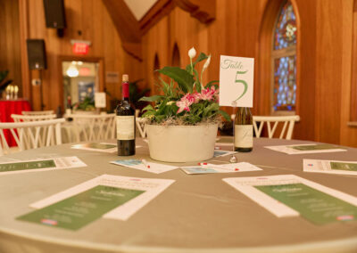 A round table is set with menus, pens, wine bottles, and a potted plant centerpiece, with a Table 5 sign. The setting is indoors with wooden walls and stained glass windows in the background.