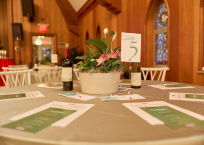 A round table with a gray tablecloth is set with menus, wine bottles, and a potted flower centerpiece. A sign reading Table 5 is in the centerpiece. The room has wooden walls and stained glass windows.