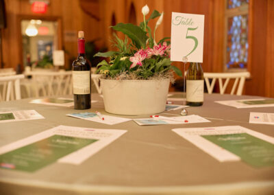 A round table with a potted plant centerpiece, two wine bottles, scattered papers, and a Table 5 sign in an indoor setting with wooden walls and white chairs.