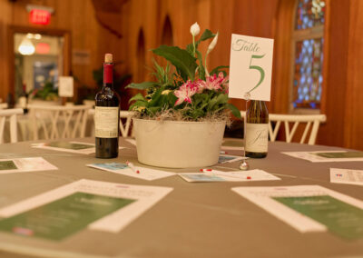 A round table set with papers, a potted plant centerpiece, a Table 5 sign, a bottle of red wine, and a bottle of white wine in a warmly lit room with wooden walls and white chairs.