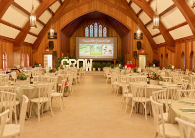 A large, elegant banquet hall with wooden arches, round tables covered in white cloths, floral centerpieces, and a stage with a screen displaying Womens Club Garden Party. White GROW letters are set near the stage.