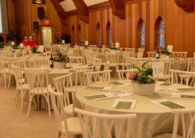 A banquet hall with round tables covered in light-colored tablecloths, each table set with papers and a potted plant centerpiece. White chairs surround the tables, and the room features wooden walls and stained glass windows.