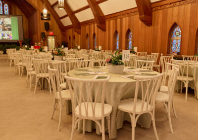 Round tables with white chairs are arranged in a spacious, wood-paneled banquet hall. Each table has a centerpiece with flowers and a table number. Stained glass windows and a large screen are visible in the background.