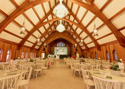 An elegant banquet hall with a high vaulted wooden ceiling, chandeliers, and round tables set for an event. A large screen and GROW letters are on stage, with stained-glass windows along the walls.