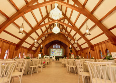 Elegant event hall with wooden arched beams, chandeliers, and lanterns. Rows of white chairs and tables line the space, facing a large screen displaying photos at the front. The ambiance is warm and inviting.
