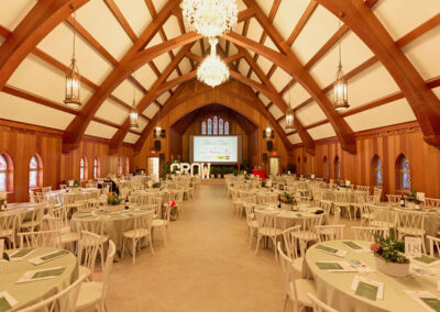 A large, elegant event hall with wooden beams, chandeliers, round tables with white chairs, and a large screen at the front displaying “GROW.” Potted plants serve as centerpieces on each table.
