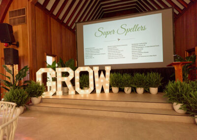An event stage features large illuminated letters spelling GROW, surrounded by potted plants. Behind the stage, a screen displays a Super Spellers list of names in a wooden-paneled room with high, vaulted ceilings.