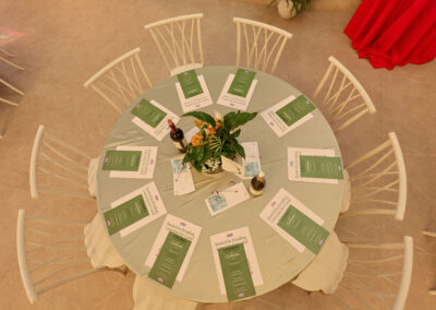 An overhead view of a round table set for eight, with green menus, bottles of wine, water glasses, and a potted plant centerpiece on a pale tablecloth. White chairs surround the table.