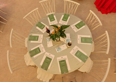 Overhead view of a round table set for eight, with white chairs, green menus, white place cards, two wine bottles, water glasses, and a potted plant centerpiece on a light tablecloth.