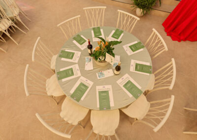 A round table set for eight with white chairs, green menus, wine bottles, water bottles, potted plant centerpiece, and a few booklets, viewed from above on a light-colored floor.