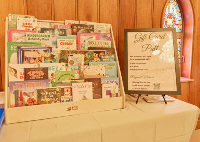A tabletop display of colorful children’s books sits next to a framed sign describing a gift card pull fundraiser, against a wooden wall with a stained glass window in the background.