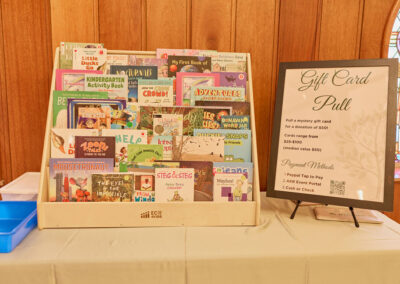 A wooden display holds various colorful childrens books on a table beside a sign titled Gift Card Pull, explaining a fundraiser with payment options, against a wood-paneled wall with stained-glass windows in the background.