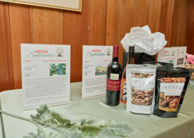 A display table with auction items including bottles of wine, two bags of Wicked Mix snacks, and printed descriptions of the items, set against a wooden wall background.