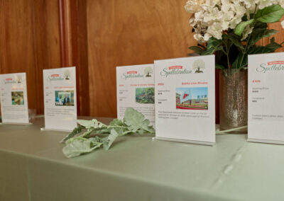 Four informational display cards are arranged on a green tablecloth beside a floral arrangement in a vase, likely part of an event or auction. The background features wooden paneling.