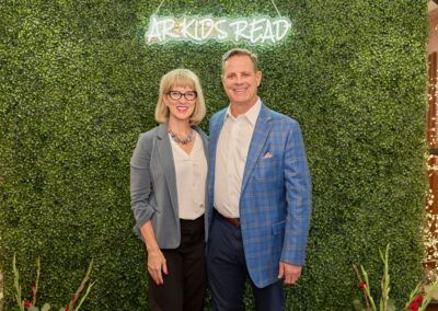 A woman and a man stand smiling in front of a green hedge wall with a neon sign that reads AR KIDS READ, surrounded by string lights and decorative plants.