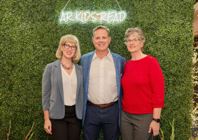 Three adults stand smiling in front of a green leafy wall with a neon sign that reads AR KIDS READ. Two women flank a man in the center; one wears a blue blazer and glasses, the other a red sweater.
