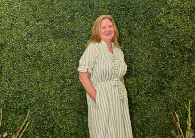 A smiling woman in a green and white striped dress stands in front of a leafy green wall with a neon sign that reads “AR KIDS READ.” Decorative plants and watering cans are placed at the bottom of the wall.