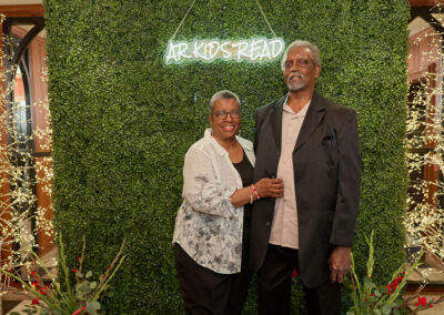An older couple stands smiling in front of a green foliage wall with a neon AR Kids Read sign. They are dressed semi-formally and are surrounded by decorative lights and plants.
