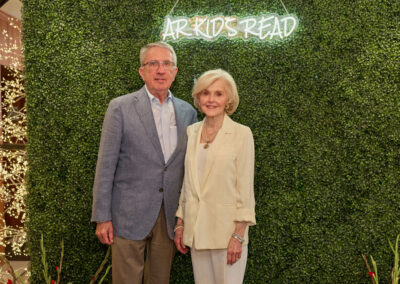 An older man and woman stand together, dressed in light-colored suits, in front of a green hedge wall with a glowing AR KIDS READ sign. Decorative lights are visible in the background.