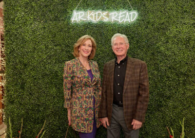 A smiling woman and man stand in front of a green leafy wall with a neon sign that reads AR Kids Read. The woman wears a patterned jacket and purple dress; the man wears glasses and a plaid jacket with a black shirt.