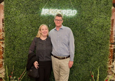 A smiling couple poses in front of a green hedge wall with a neon sign that reads ARKIDS READ. The woman holds a jacket and a purse, and both are dressed in business-casual attire. Decorative lights and flowers are in the foreground.
