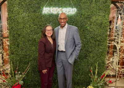 A woman and a man stand smiling in front of a green hedge wall with a neon ARKIDS READ sign. They are dressed in business attire and surrounded by decorative lights and red flowers.
