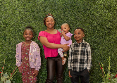 A woman stands smiling with three children in front of a green leafy backdrop and a neon sign that reads AR KIDS READ. Two children stand on either side, and she holds a baby. Colorful watering cans and flowers are on the floor.