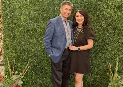 A man in a blue blazer and a woman in a black dress stand smiling together in front of a green leafy wall with a neon sign that reads AR KIDS READ. Decorative plants and watering cans are on either side of them.