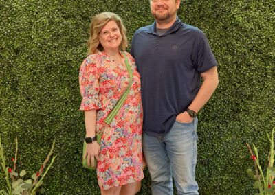 A woman in a floral dress and a man in a navy shirt and jeans stand smiling together in front of a green leafy backdrop with a neon AR KIDS READ sign above them. Decorative plants and watering cans are on the floor nearby.