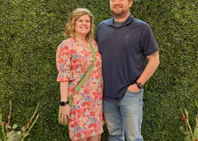 A smiling woman in a floral dress and a man in a navy polo and jeans stand together in front of a green plant wall with a neon sign that reads “AR KIDS READ.” Colorful watering cans and plants are at their feet.