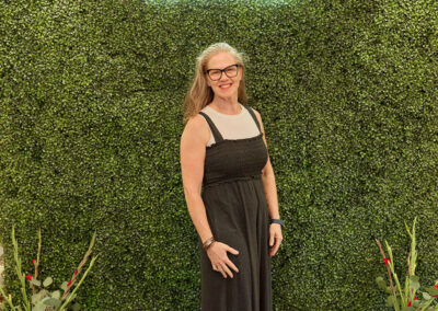 A woman in a black dress and glasses stands smiling in front of a green leafy wall with a neon sign reading “AR KIDS READ.” Decorative plants and colorful watering cans are placed on the floor.